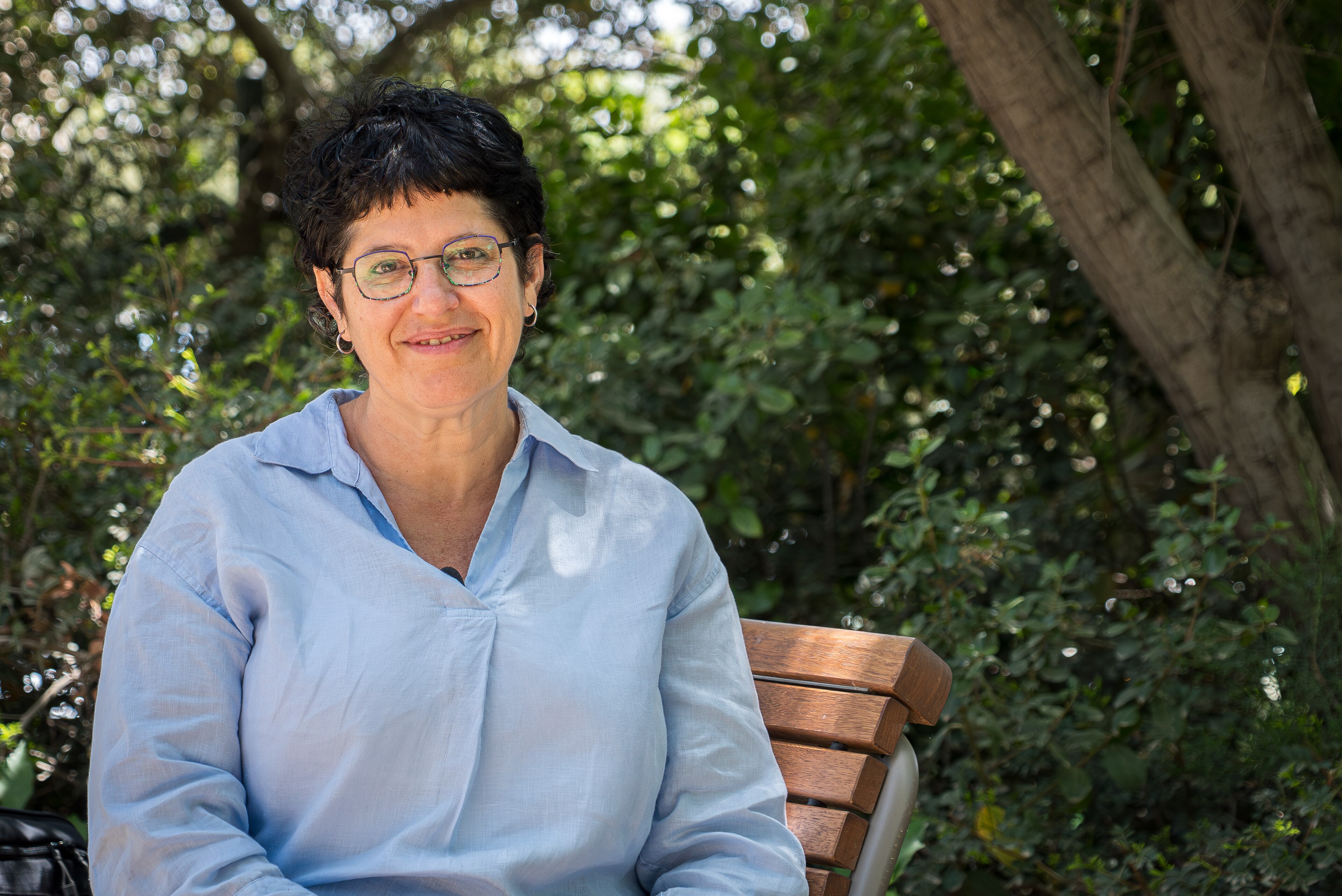 A close up of a woman with dark hair and glasses smiling as she sits.