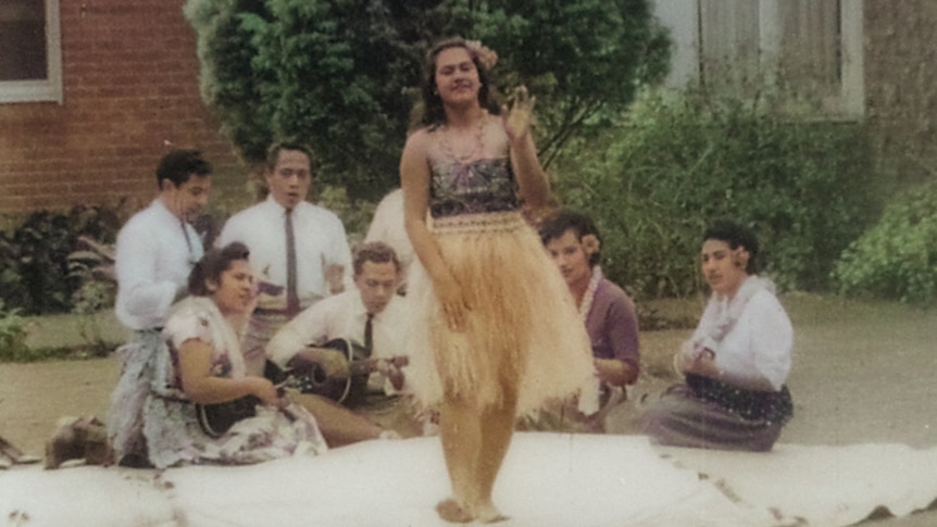 A woman dances on a woven mat while seven men and women sing and play stringed instruments behind.