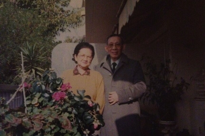 An old photo showing an older Vietnamese man and woman, standing in a garden by a house.