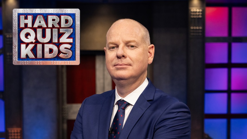 Tom in a blue suit and tie with his arms crossed. He stares at the camera. The Hard Quiz Kids logo is in the top left corner