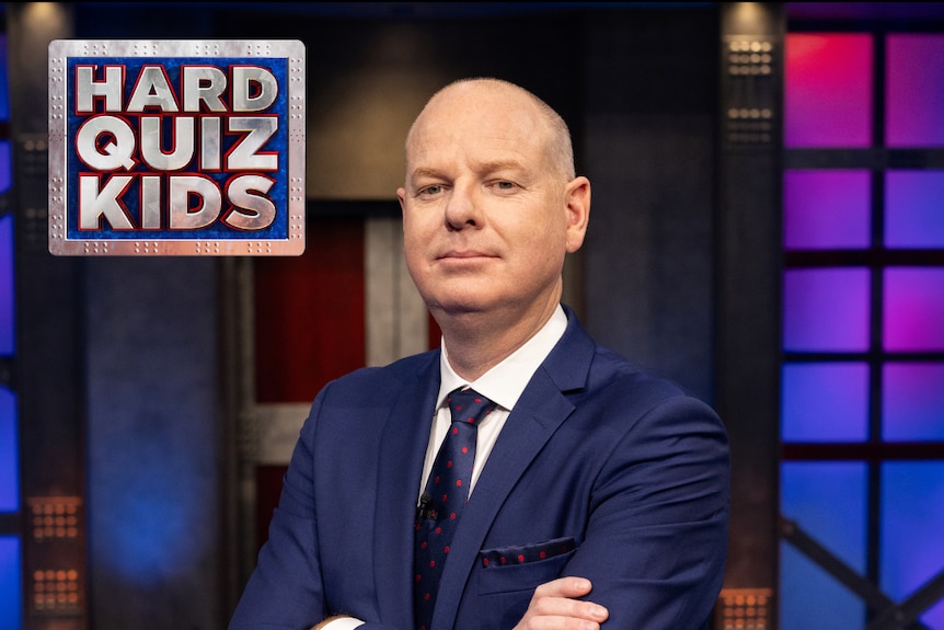 Tom in a blue suit and tie with his arms crossed. He stares at the camera. The Hard Quiz Kids logo is in the top left corner