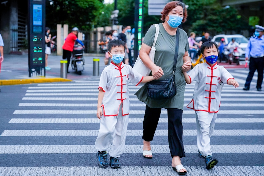 An older Chinese woman crosses a street holding the hands of twin boys in masks 