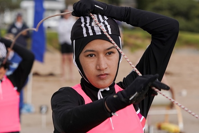 Sana Mosawi, wearing a black and pink wetsuit, reels a rope over her head on a Victorian beach.