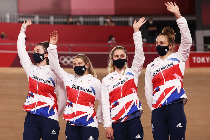 Four women cyclists from Great Britain wear masks and stand on the podium waving their arms