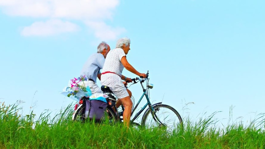 Picture of couple riding bikes. There is blue sky behind them and they are riding over a green hill 