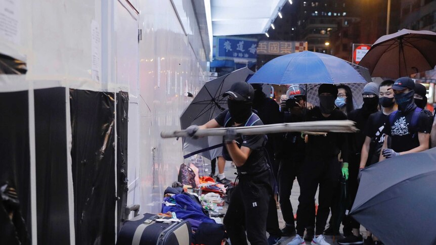 Protesters in black  masks and with blue umbrellas hit a Bank of China wall with a baton.