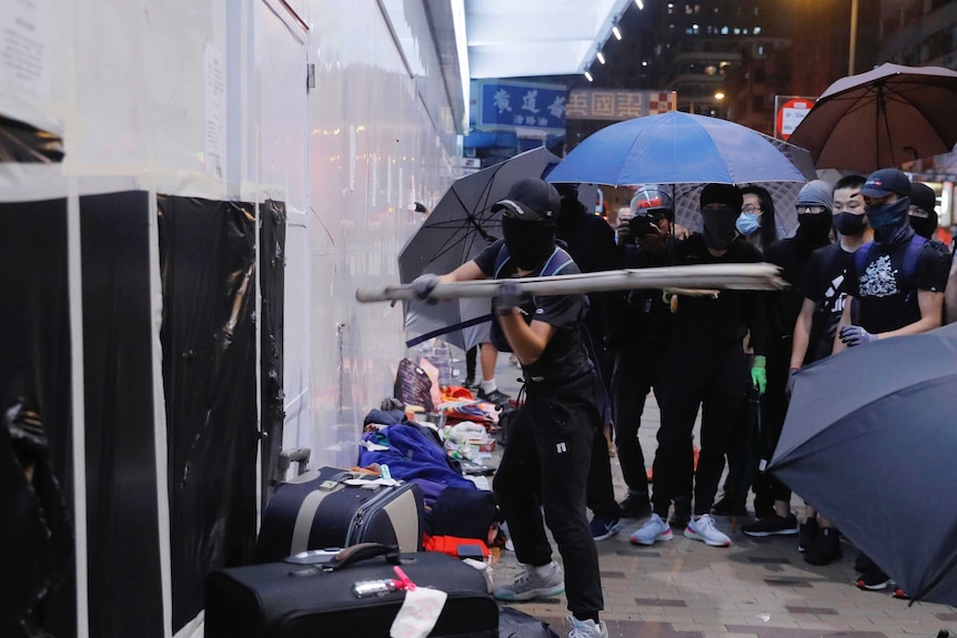 Protesters in black  masks and with blue umbrellas hit a Bank of China wall with a baton.