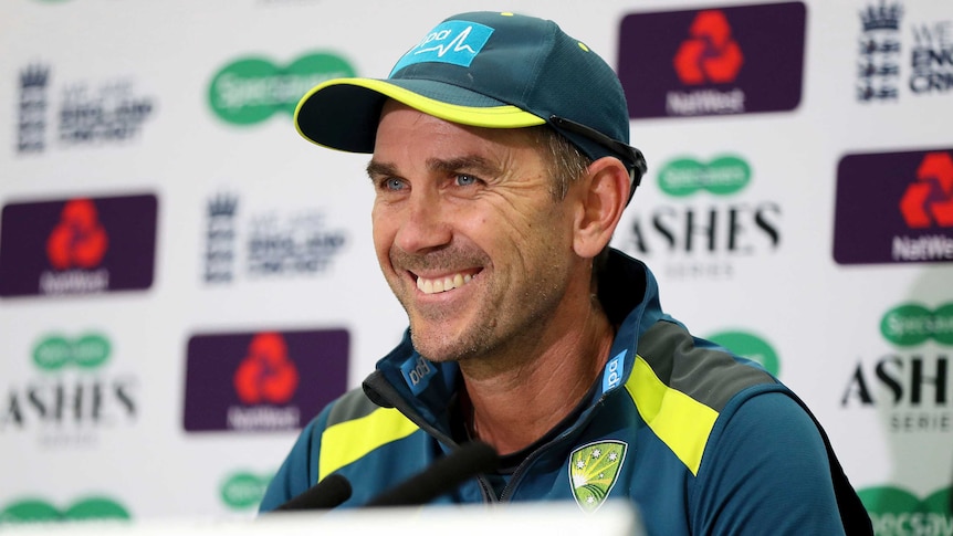 A cricket coach smiles at the media as he sits behind a desk at a press conference.