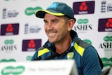 A cricket coach smiles at the media as he sits behind a desk at a press conference.
