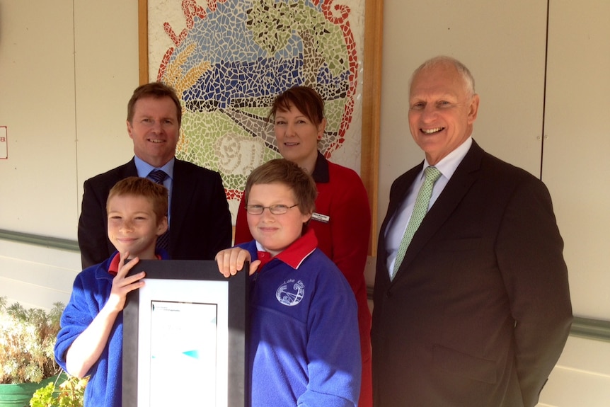 Kaye Brownley, David Axworthy, Ken Perris and two students stand in front of a mosaic school emblem and hold a certificate