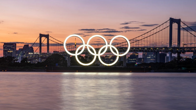 At sunset, you view illuminated white Olympic rings over a still Tokyo Bay, with a two-pylon suspension bridge behind it.