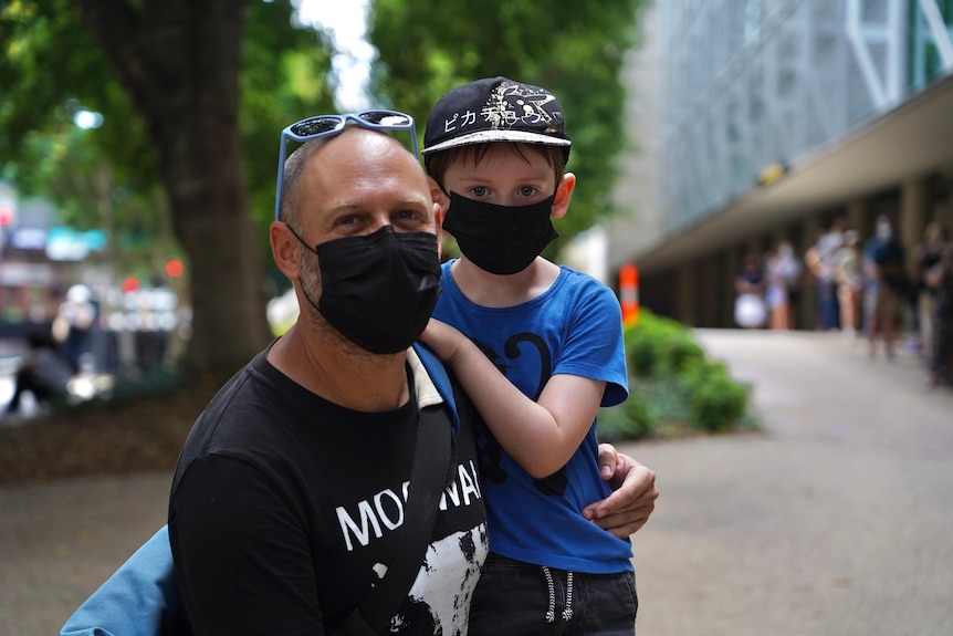 A bald man with a black t-shirt and sunnies on his head holds his young son, wearing a blue t-shirt and black cap, on his lap