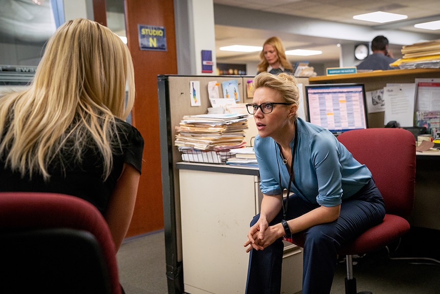 A seated woman in office cubicle with serious expression, blonde hair and black spectacles leans towards female coworker.