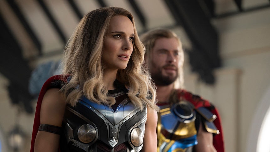 White woman with curled blonde hair wears blue chrome armor and red cape, while blonde man wears the same in background.