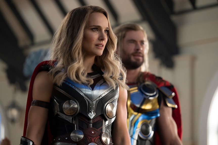 White woman with curled blonde hair wears blue chrome armor and red cape, while blonde man wears the same in background.