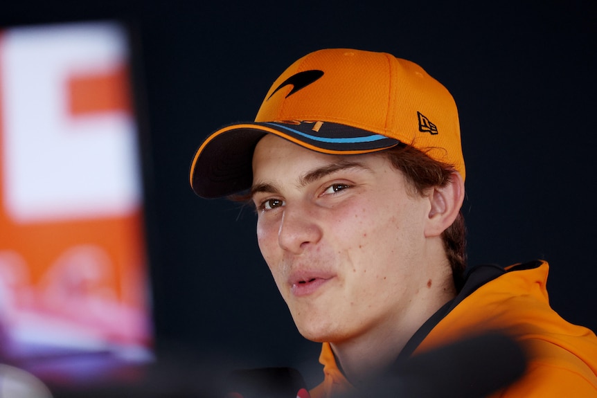 An F1 driver in an orange shirt and cap, sits in his team garage during a session