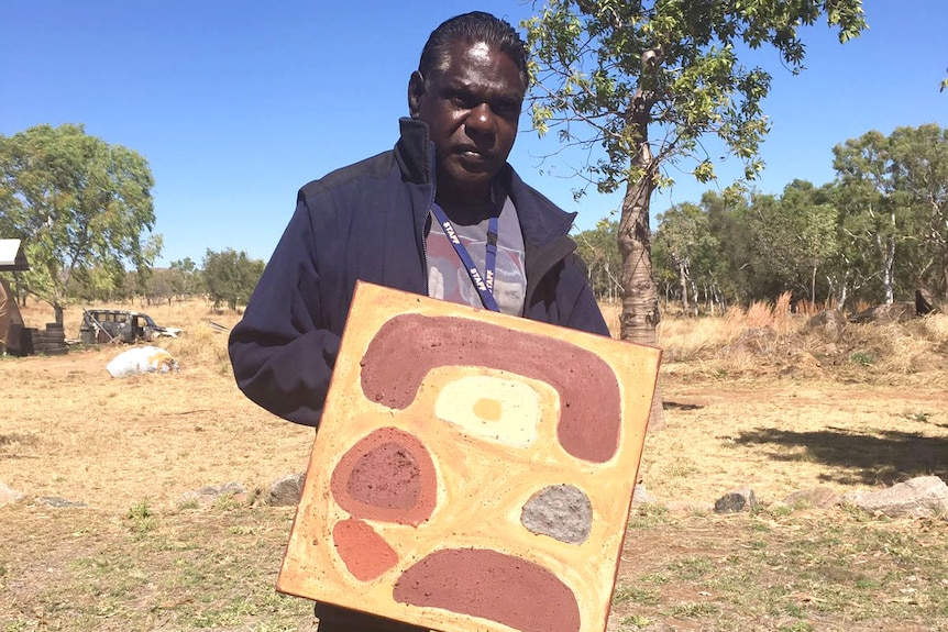 Artist Gabriel Nodea holding a dot painting while standing in bright sunlight in a bush setting.