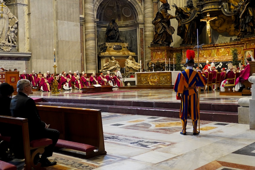 People in religious robes sit and stand in a church where a coffin lies. 