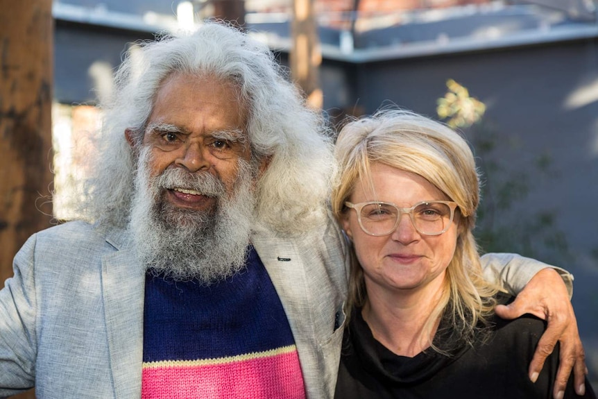 A smiling man with long white hair and a grey beard stands with his arm around a grinning blonde woman.