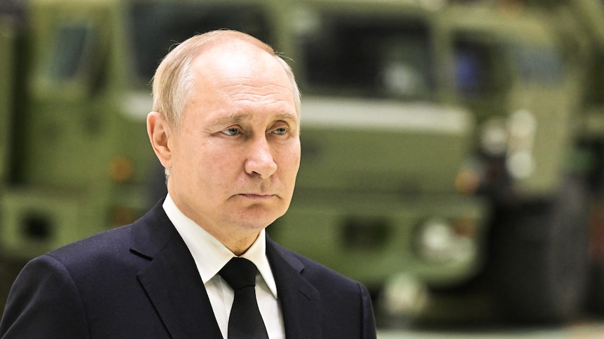 Vladimir Putin wears a suit with a black tie while standing infront of a green tank.