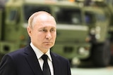 Vladimir Putin wears a suit with a black tie while standing infront of a green tank.