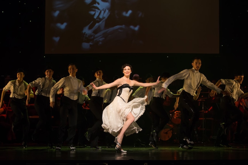 Woman in white dress, wearing silver shoes with an ensemble of male dancers. 