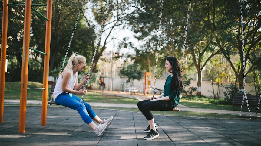 Two young women sit on a swing set facing each other, having a happy conversation.