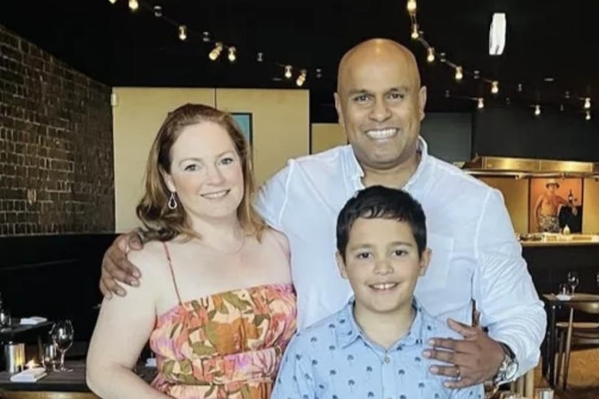 The family stands in a restaurant with Raj holding his arm around Emma's shoulder and his other hand on Arli's shoulder.