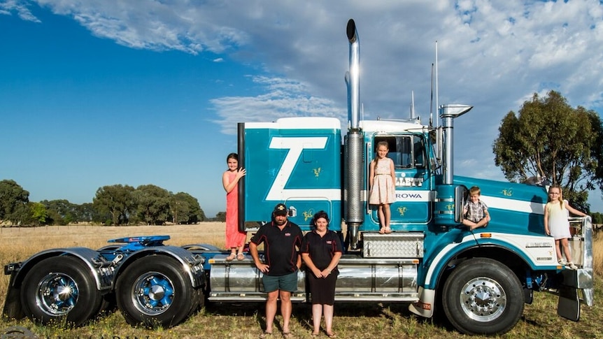 Two adults and four children stand near and on a blue prime mover truck