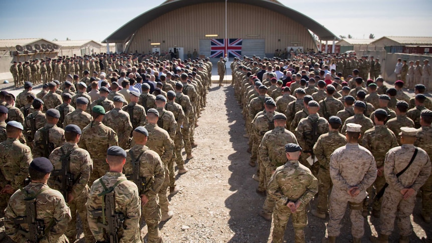 rear view of rows of troops, heads bowed, hands behind backs, in front of a British flag on a corrugated iron aeroplane hanger