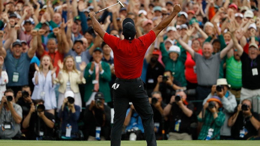 A man stands before an applauding crowd with his arms raised victoriously as he holds a golf putter in his left hand.