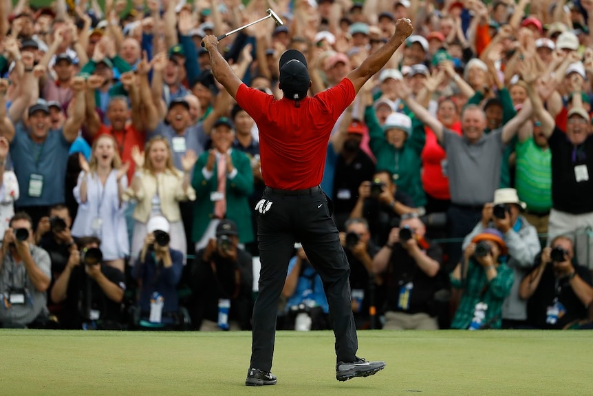 A man stands before an applauding crowd with his arms raised victoriously as he holds a golf putter in his left hand.