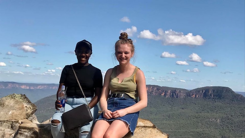 Winnie Phillips, left, in a black t-shirt and cap sits with a friend on a rock overlooking mountains.