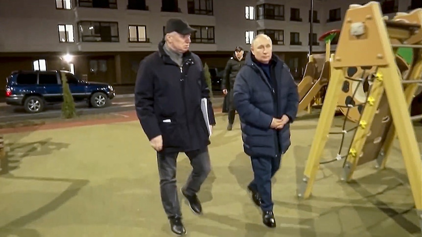 Vladimir Putin wears a winter puffer jacket as he walks along a city street at night with a man