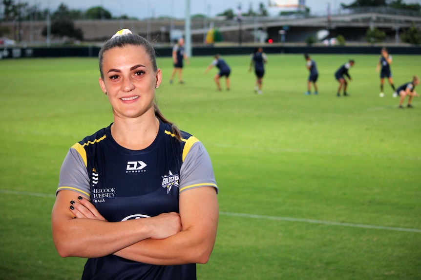 A on a football field woman smiles with her arms crossed while other players warm up in the background