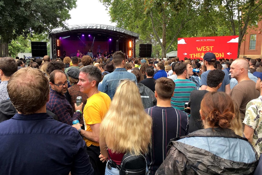 Thousands of people stand before a stage with a band playing among trees on a university ground.