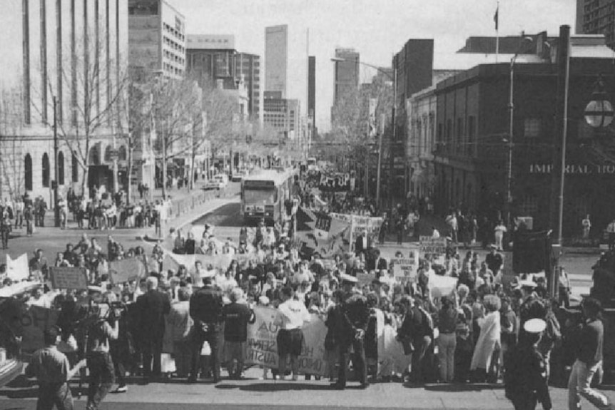 A black-and-white photograph shows a crowd of people gathered at the steps of Victorian Parliament House.