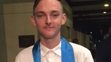 Jacob Cummins stands posing for a photo in a white shirt and black pants and with a blue satin scarf hanging around his neck.