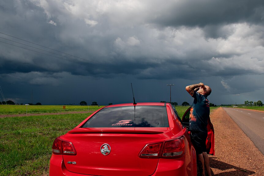 A man stands by a red car, storm clouds and a clear field in the background. 