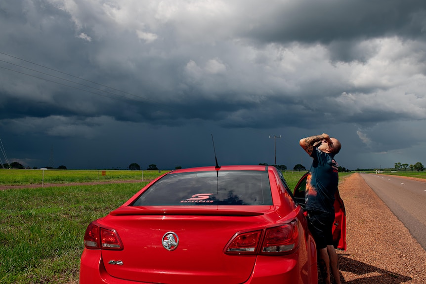 A man stands by a red car, storm clouds and a clear field in the background. 