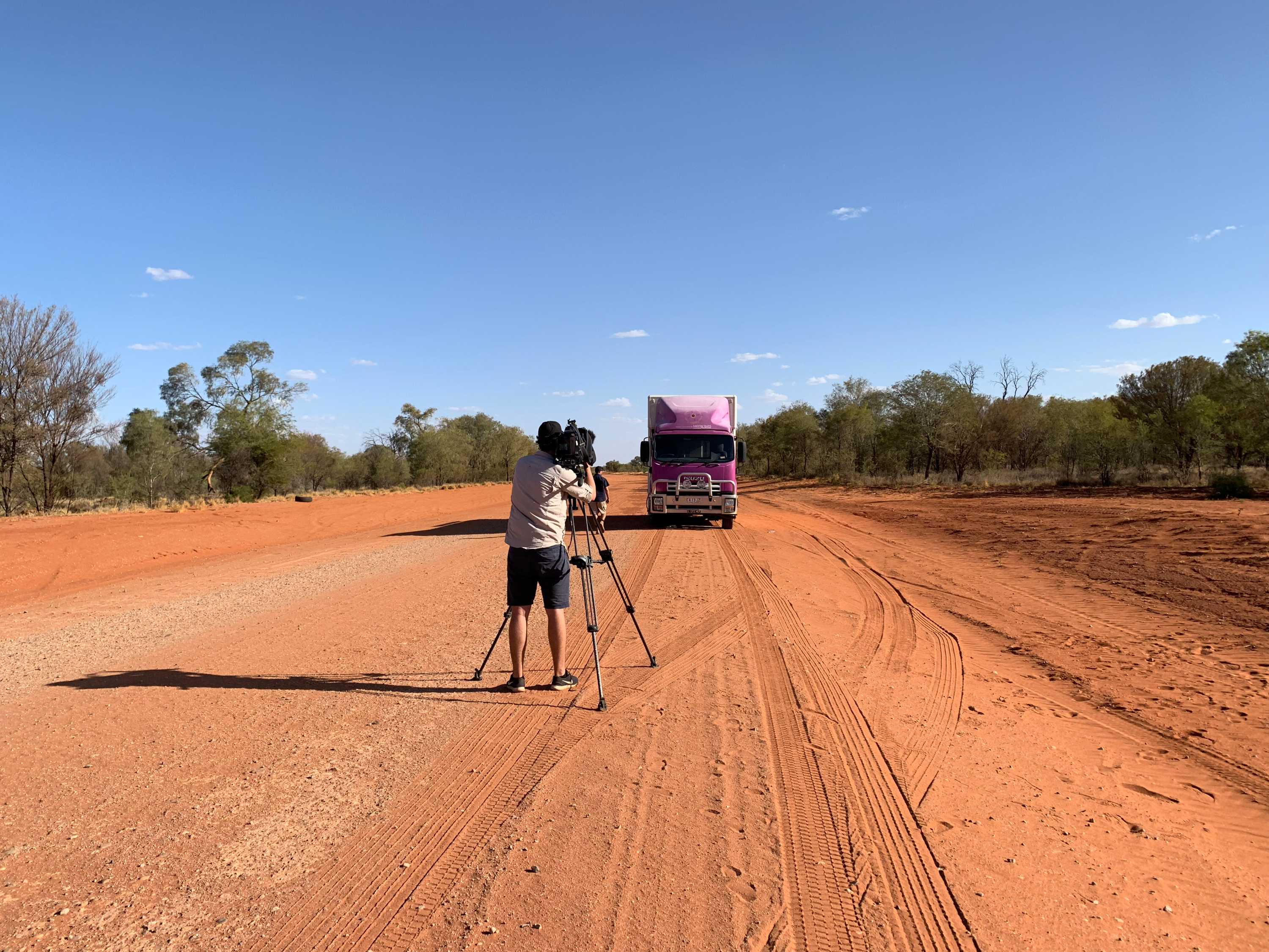 Cameraman with camera on tripod filming bus driving towards him on isolated red road.