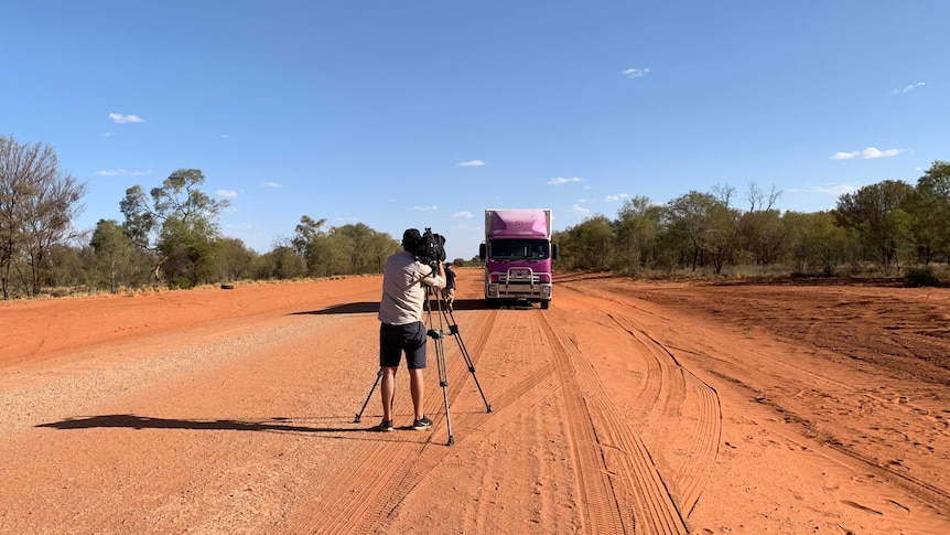 Cameraman with camera on tripod filming bus driving towards him on isolated red road.