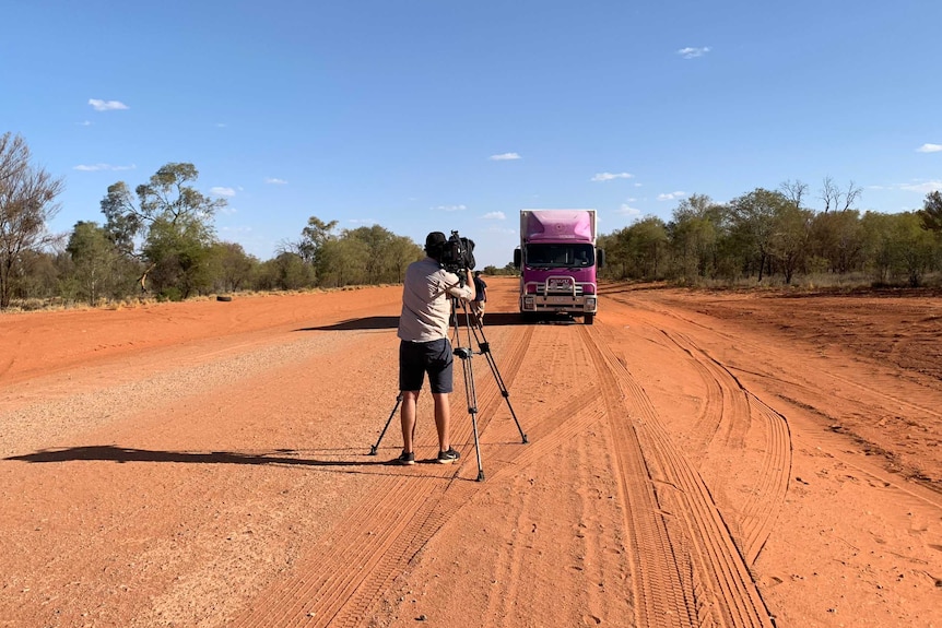 Cameraman with camera on tripod filming bus driving towards him on isolated red road.