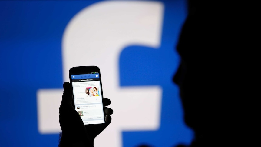 A man is silhouetted against a video screen with an Facebook logo as he poses with a smartphone.