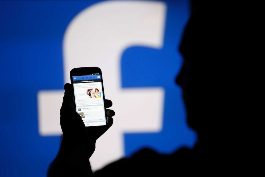 A man is silhouetted against a video screen with an Facebook logo as he poses with a smartphone.