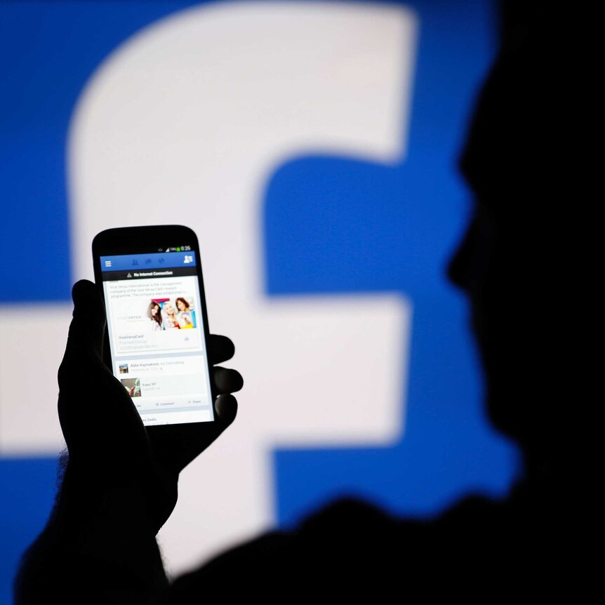 A man is silhouetted against a video screen with an Facebook logo as he poses with a smartphone