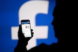 A man is silhouetted against a video screen with an Facebook logo as he poses with a smartphone.