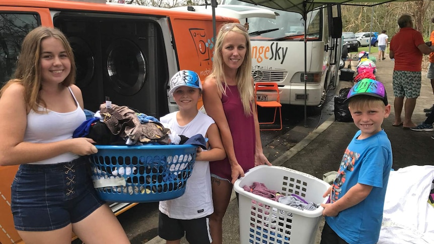 A woman with three children stand in front of an orange van holding baskets of clean clothes