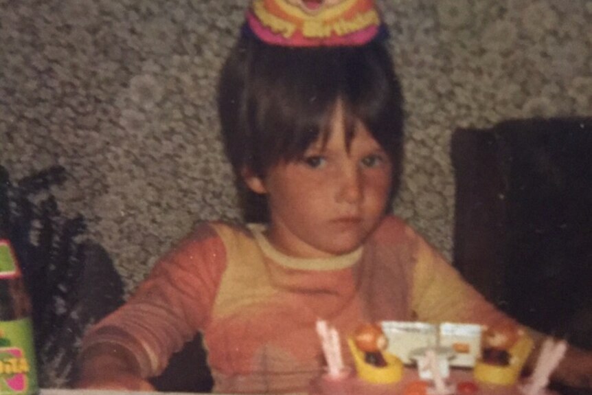 A young girl wearing a party hat sitting in front of a birthday cake. 