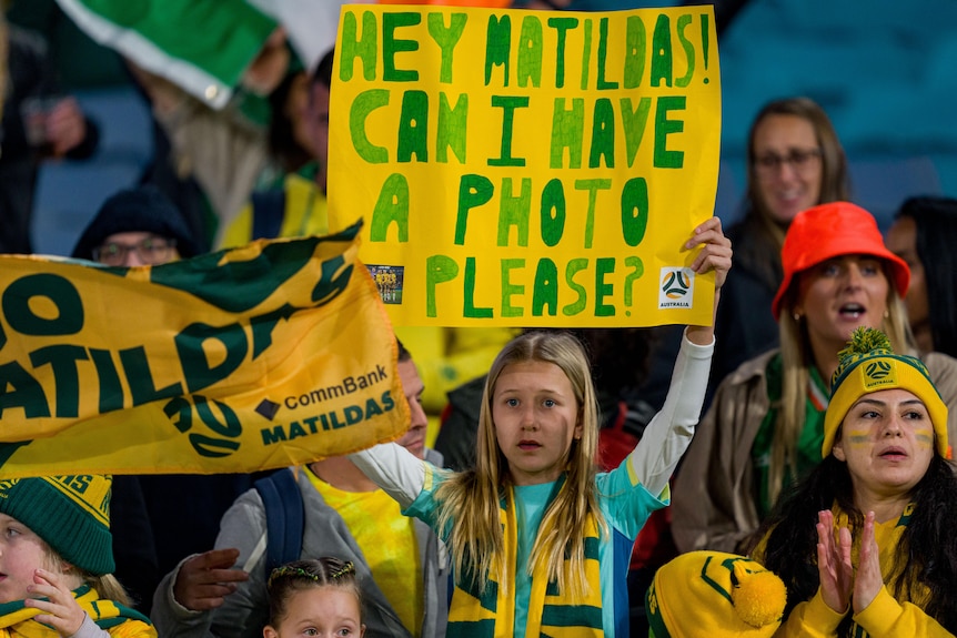 A young football fan with a sign that reads 'Hey Matildas Can I have a photo please?'